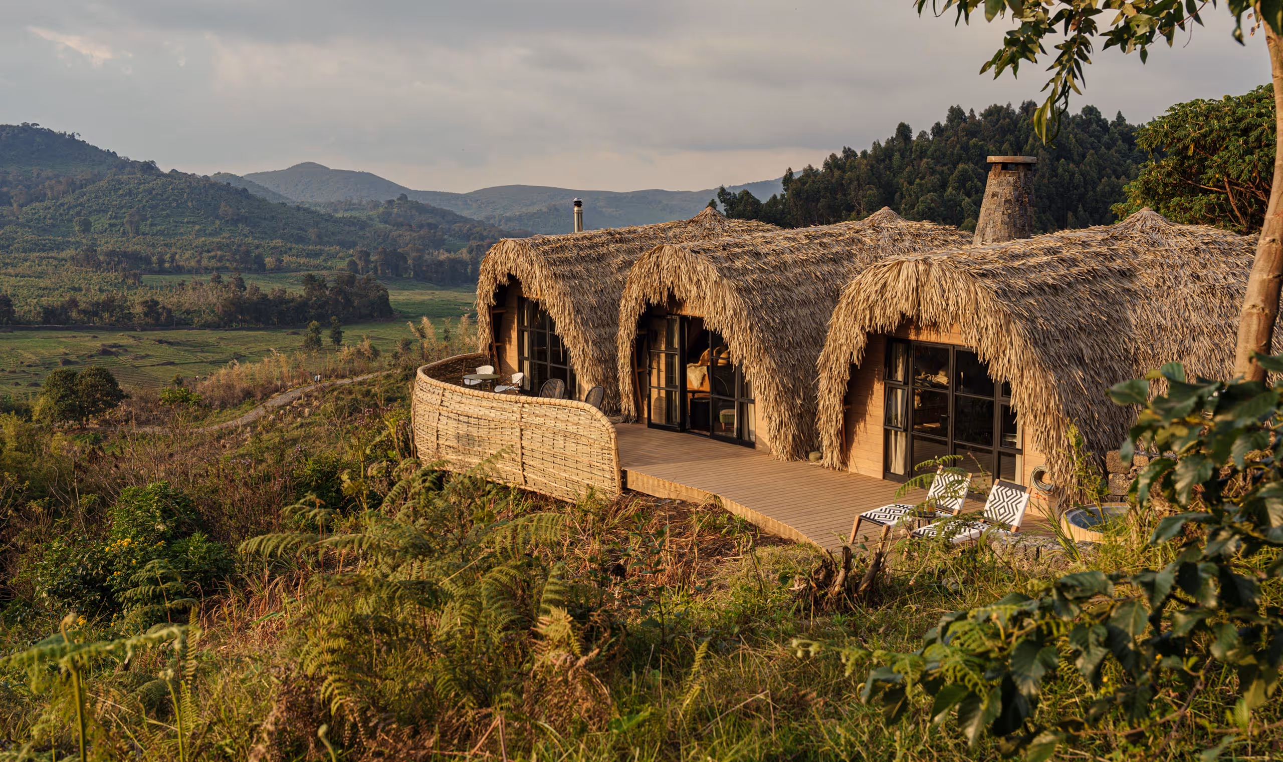 Thatched roof lodges on a hillside with mountain and forest views under a cloudy sky.