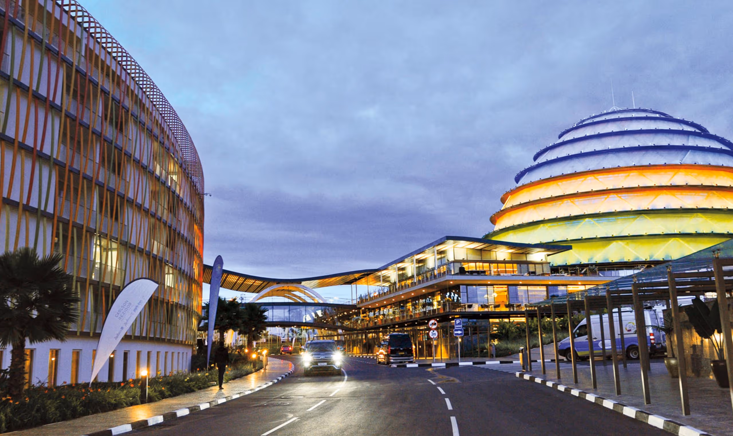 Street view of a modern building complex with a colorful dome and cars on the road at dusk.