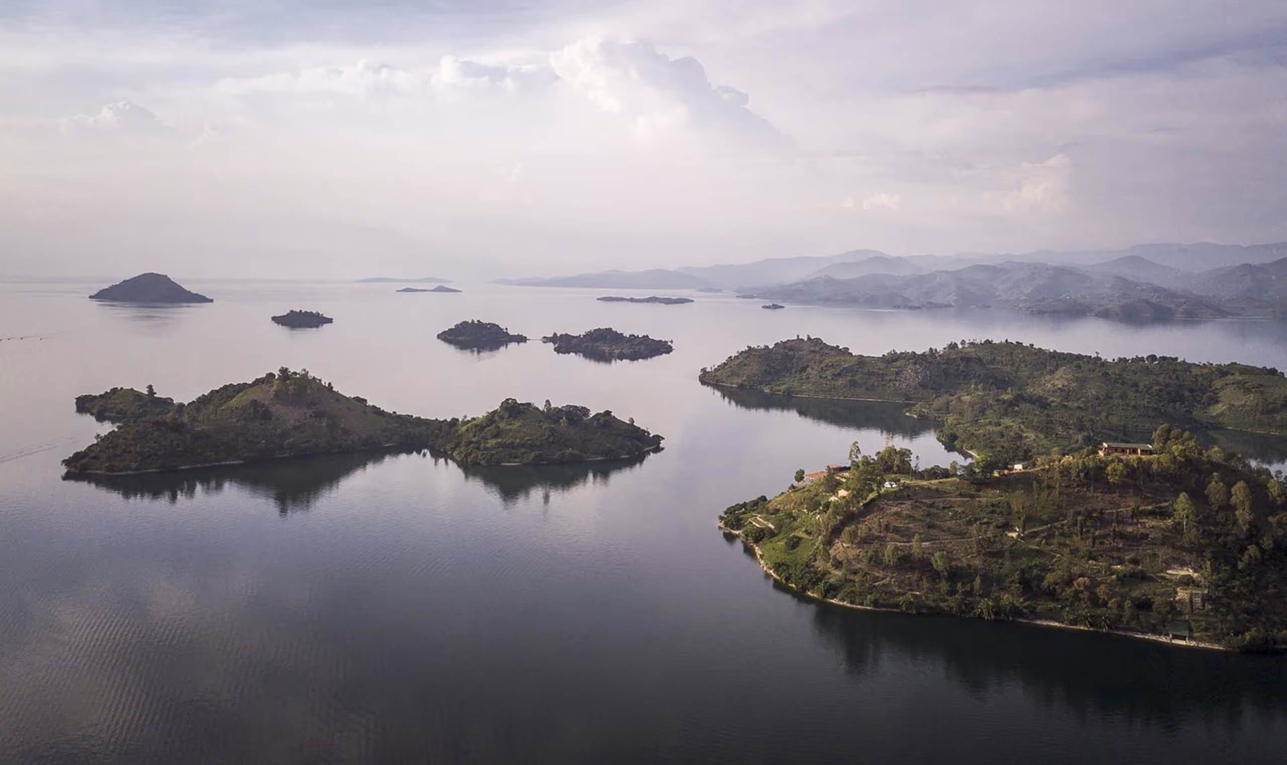 Aerial view of multiple green islands on a calm lake with hills in the distant background under a cloudy sky.