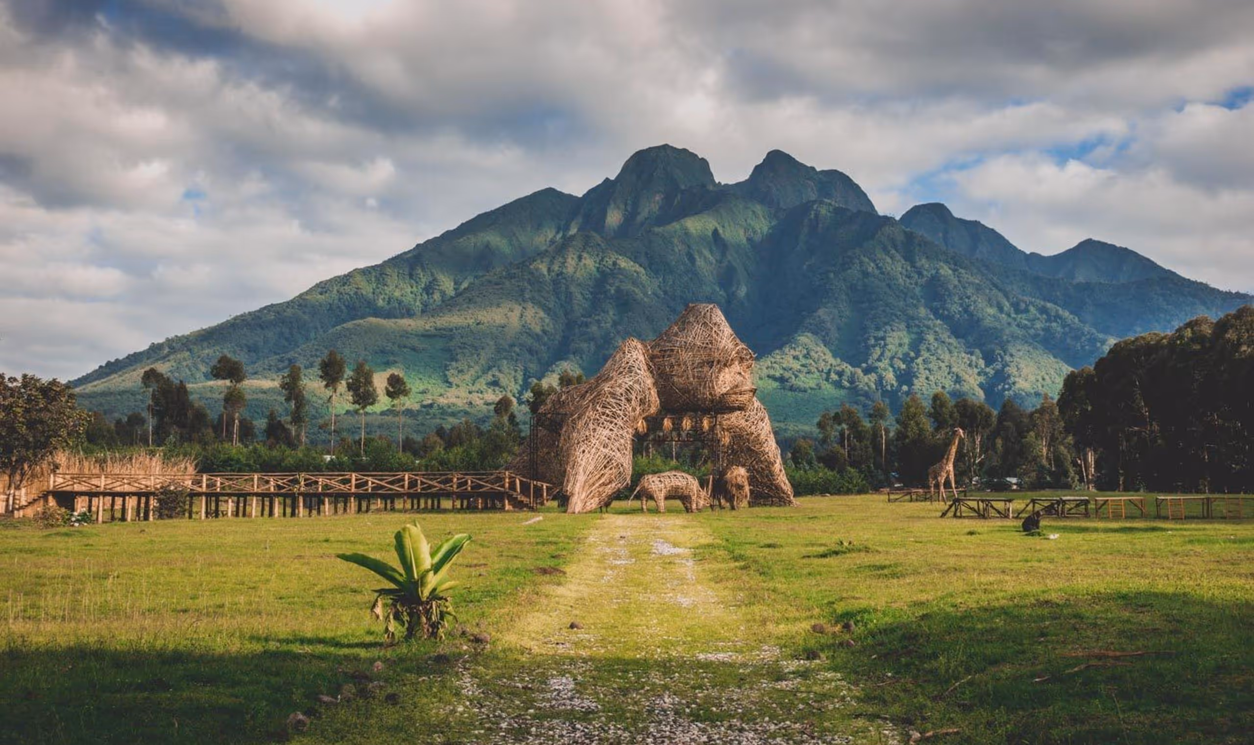 Woven gorilla sculpture and animal figures on grassy field with mountain backdrop under cloudy sky.