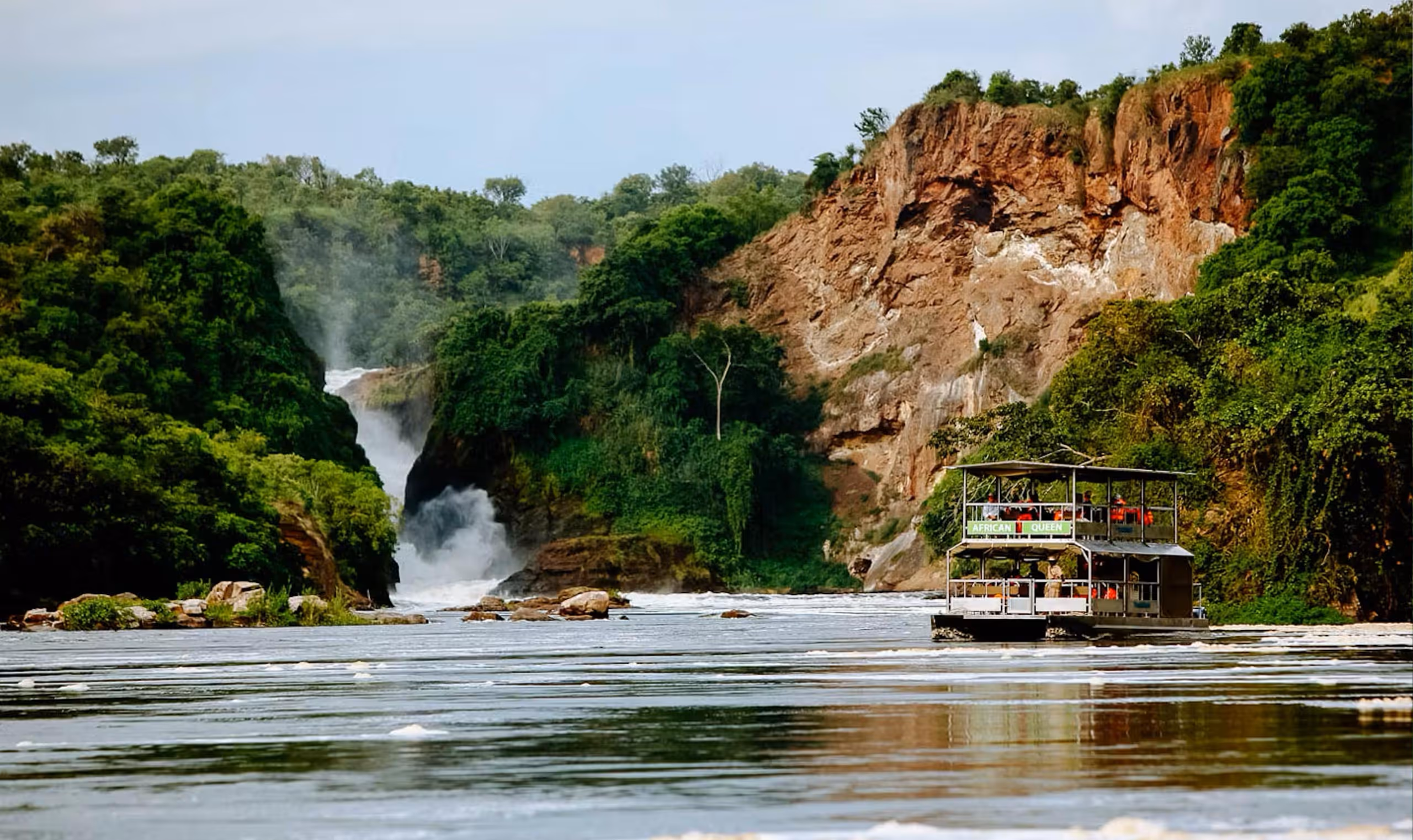 Two-level boat named African Queen on a river near rocky cliffs and a waterfall surrounded by green vegetation.
