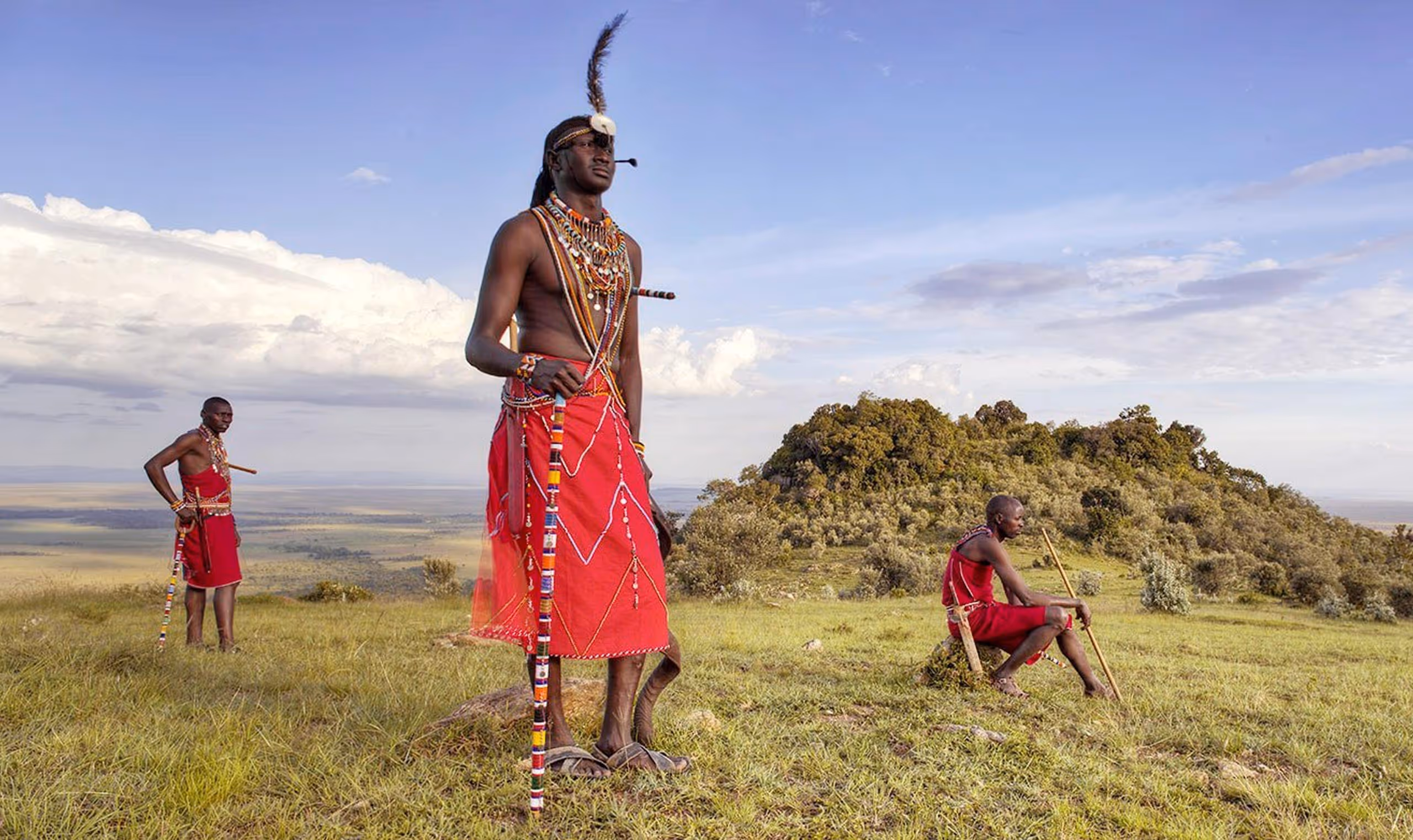 Three Maasai men in traditional red attire with beaded accessories standing and sitting on grassy savanna.