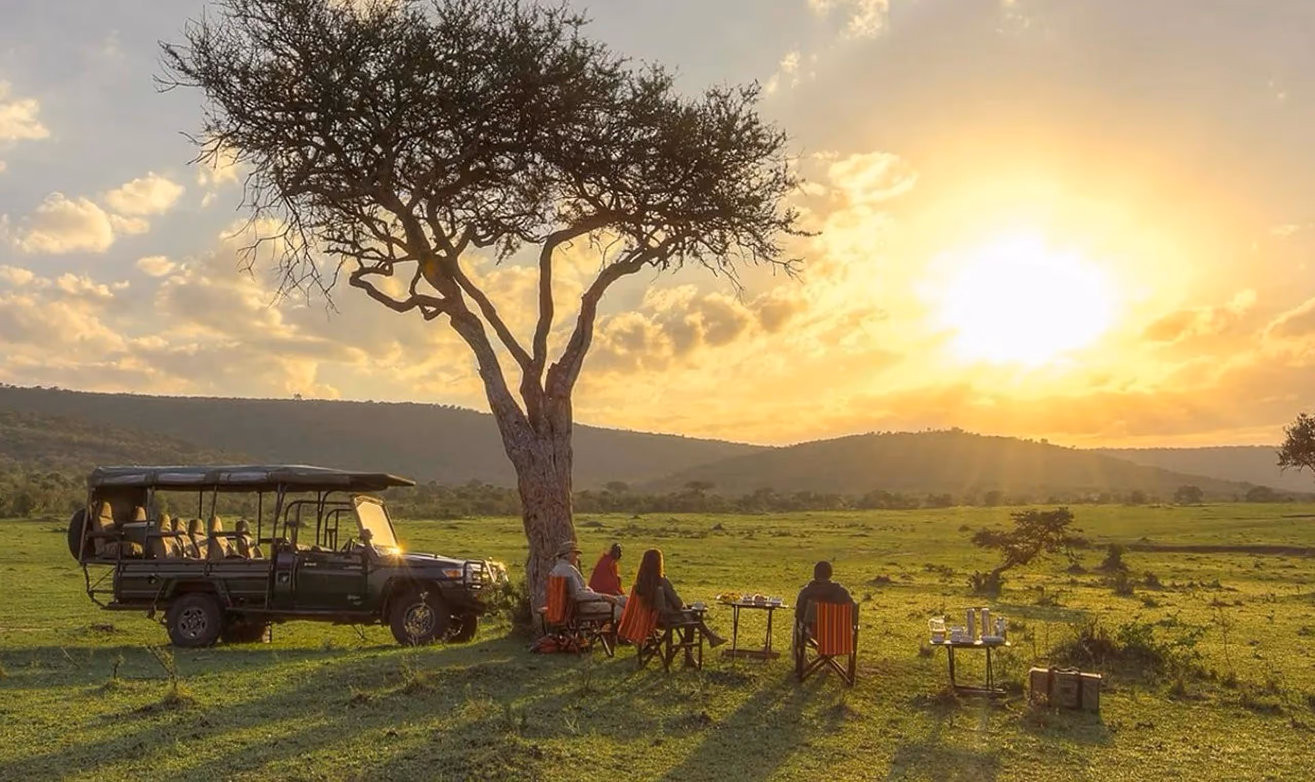Safari vehicle and four people sitting in chairs under a tree at sunset in a grassy savanna.