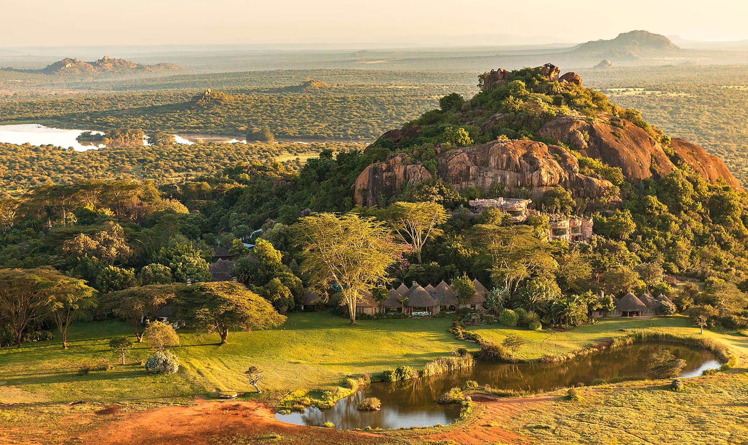 Lush green landscape with rocky hill, scattered trees, waterbody and thatched huts at its base.