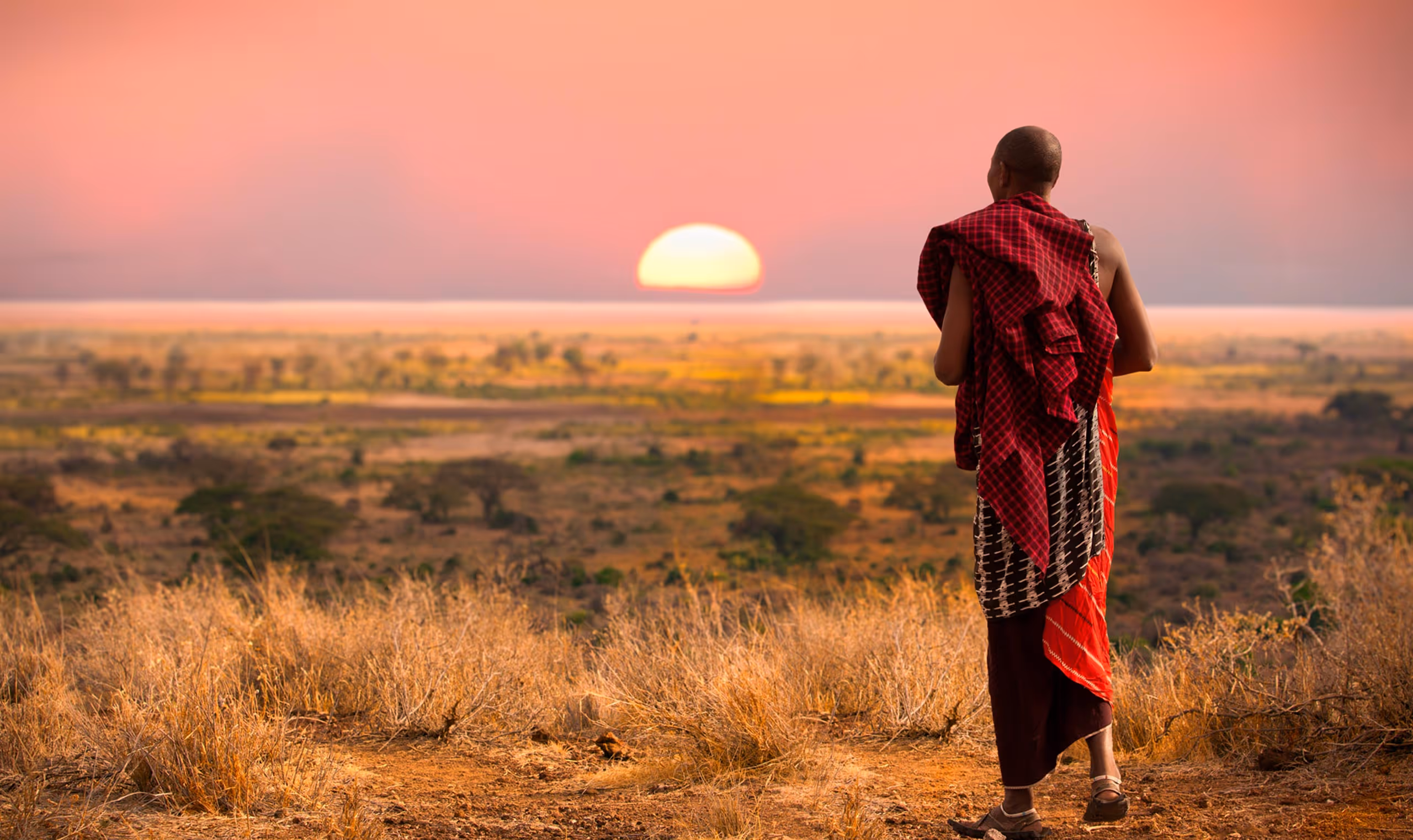 Person in traditional red Maasai attire watching the sun set over a savannah landscape.