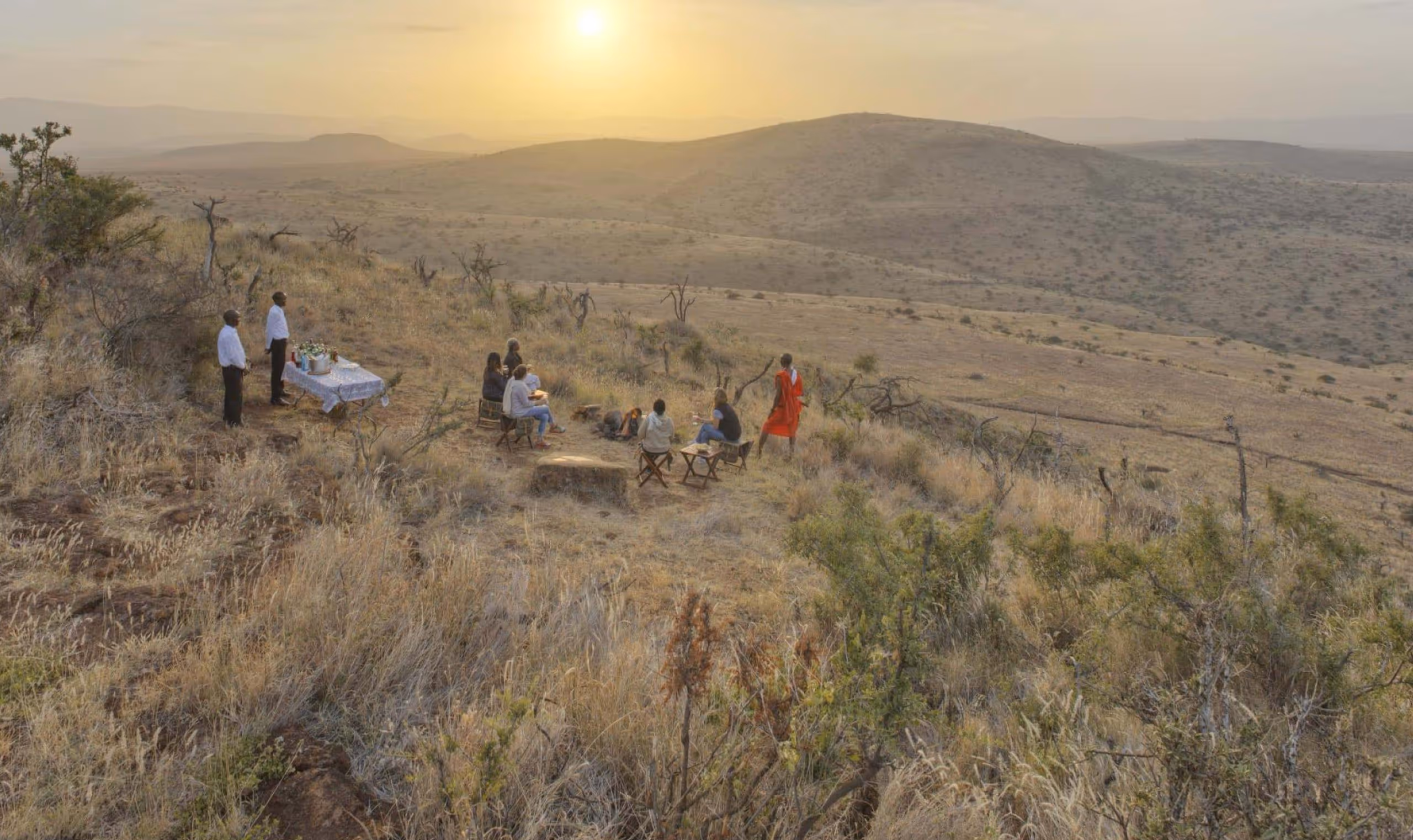 Group seated outdoors on dry hills at sunset with two men standing by a table with drinks and flowers.