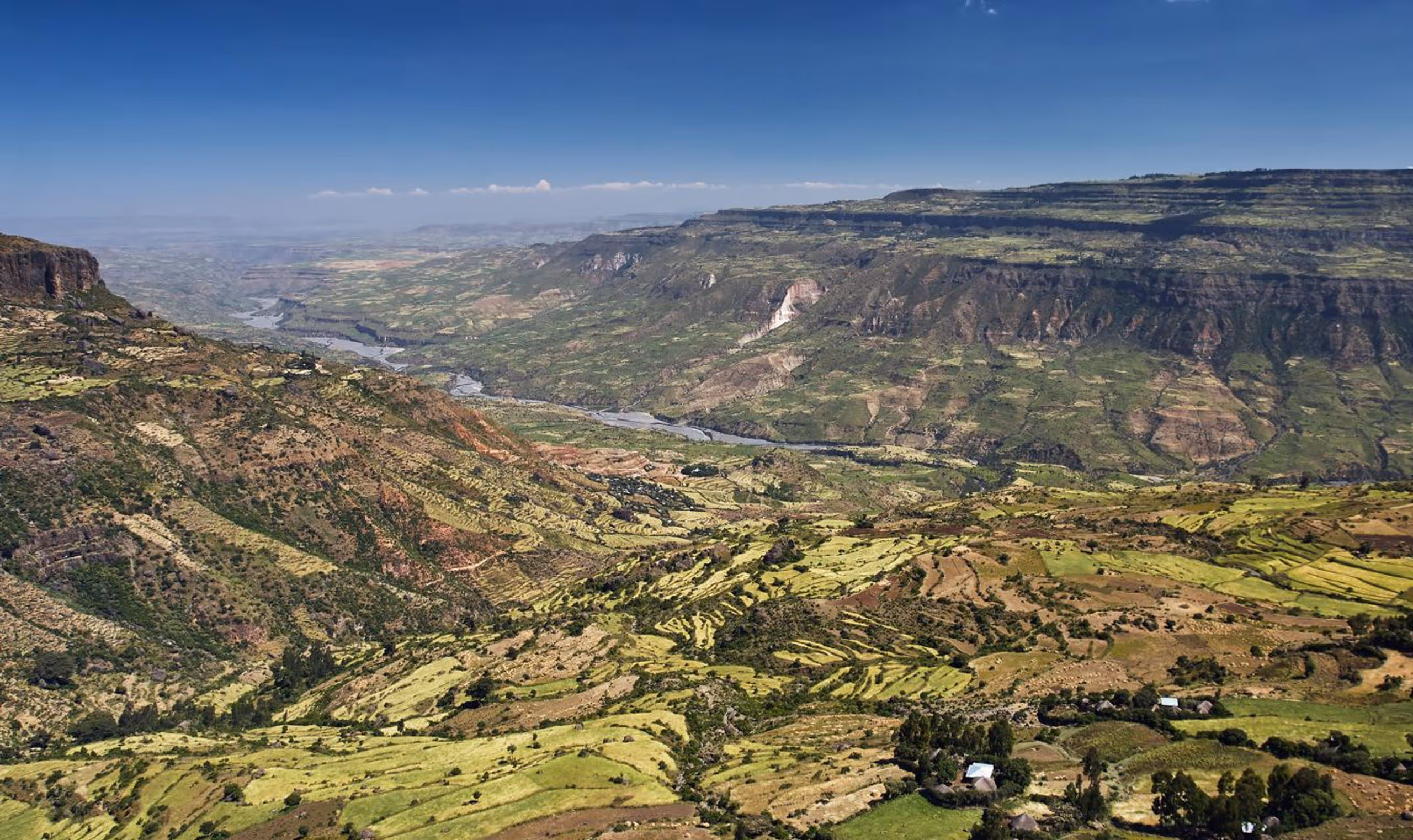 Vast valley with terraced green fields, a winding river, and layered cliffs under a clear blue sky.