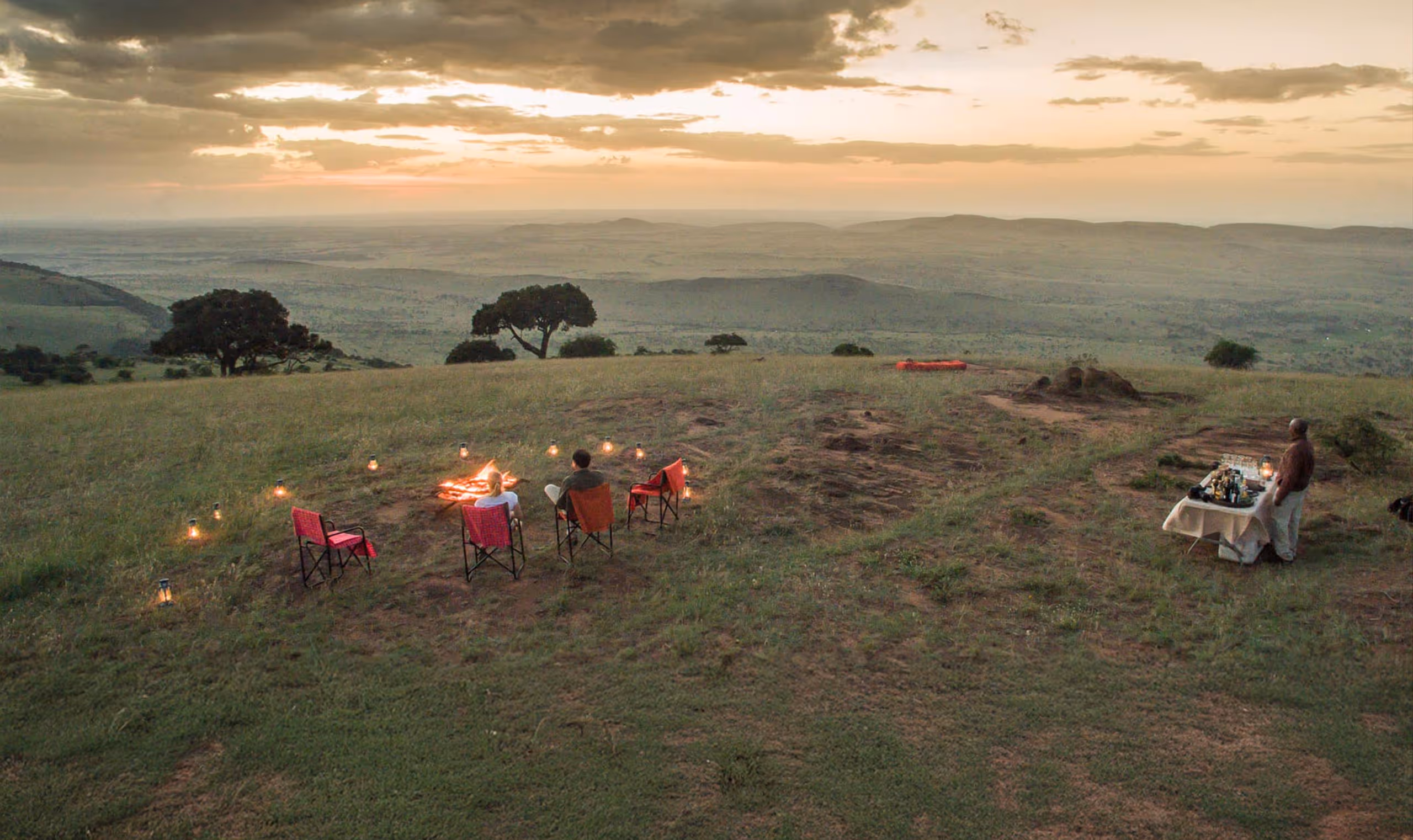 Two people sit by a campfire with lanterns on a grassy hill at sunset, overlooking distant hills.