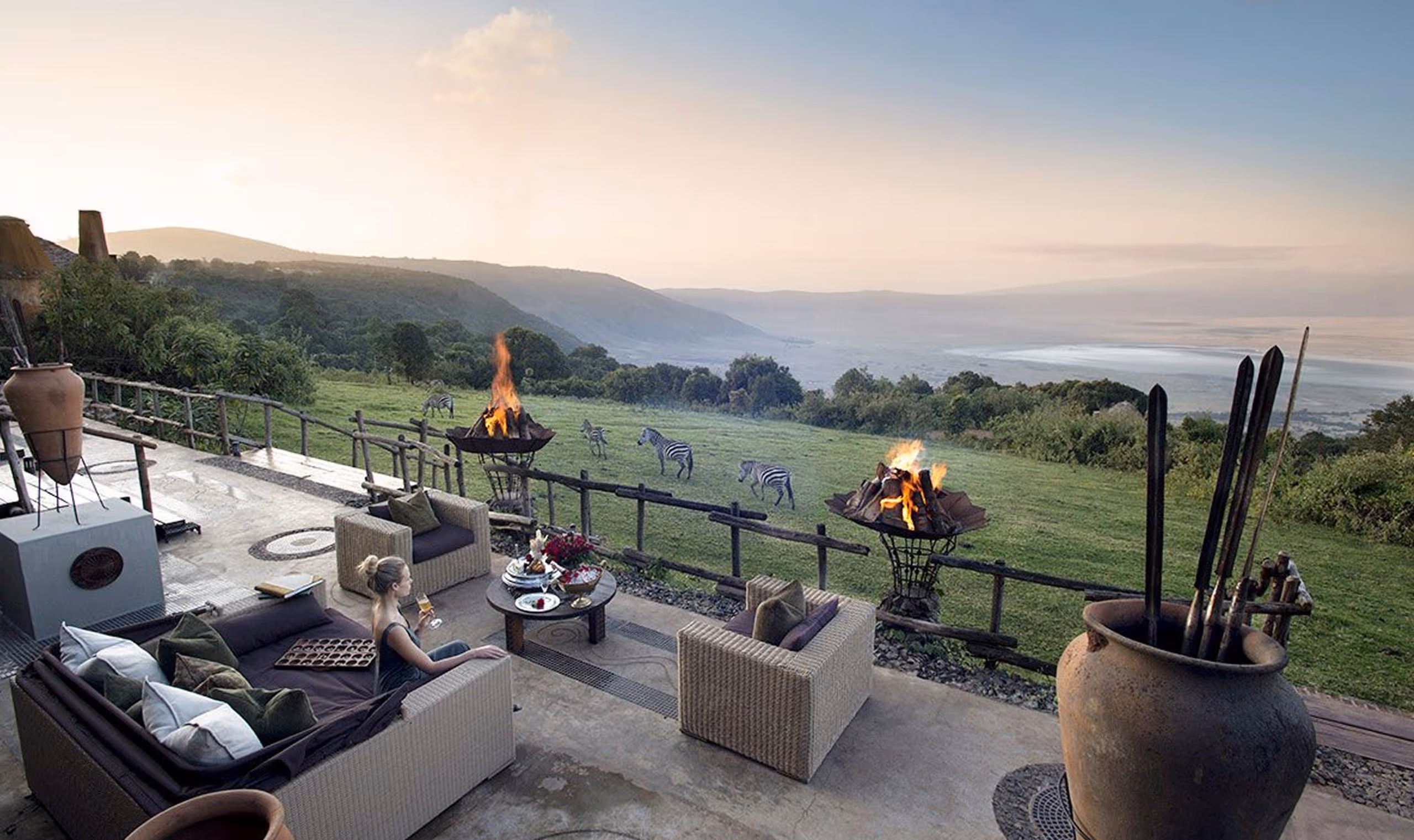 Woman sitting on a patio with fire pits, overlooking zebras grazing and a vast valley at sunset.