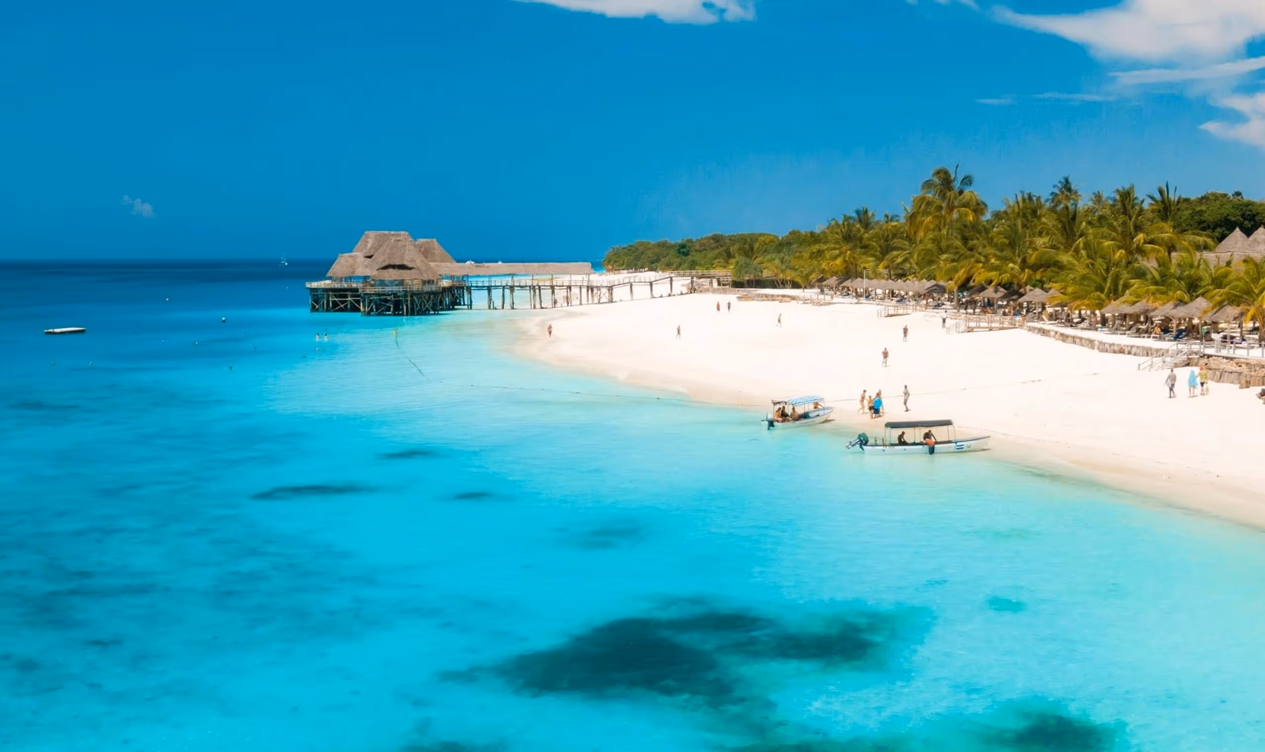 White sandy beach with clear turquoise water, palm trees, boats, people, and a wooden pier with thatched huts.