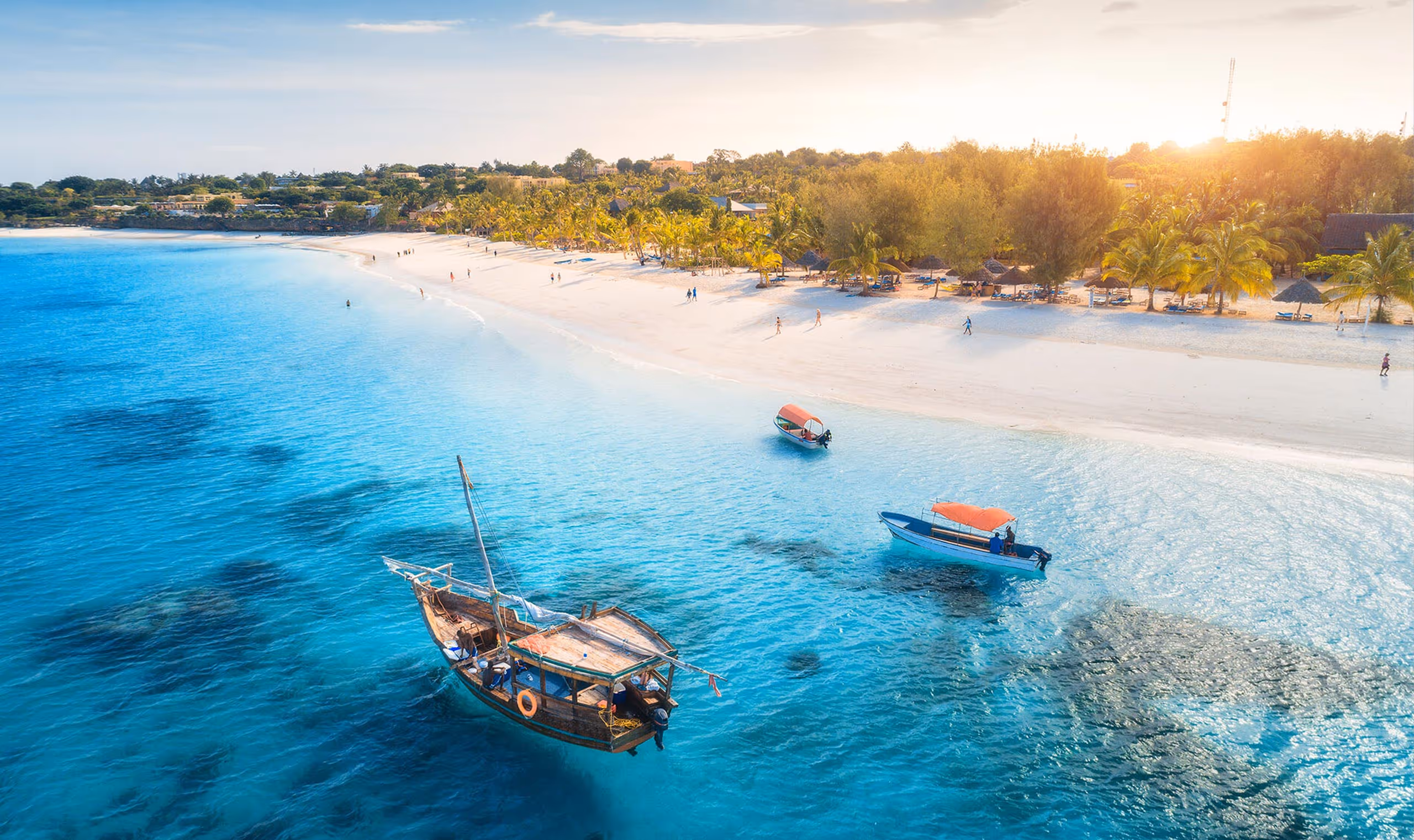 Three boats floating on clear blue water near a white sandy beach lined with palm trees at sunset.