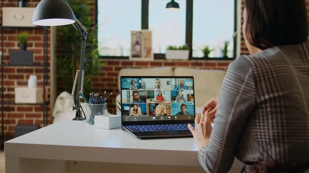 Person sitting at a desk with a laptop showing a video conference call with nine participants in a brick-walled room.