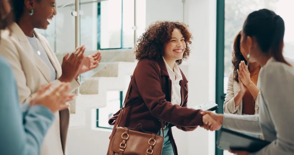 Smiling woman shaking hands with another woman while others applaud in a modern office setting.