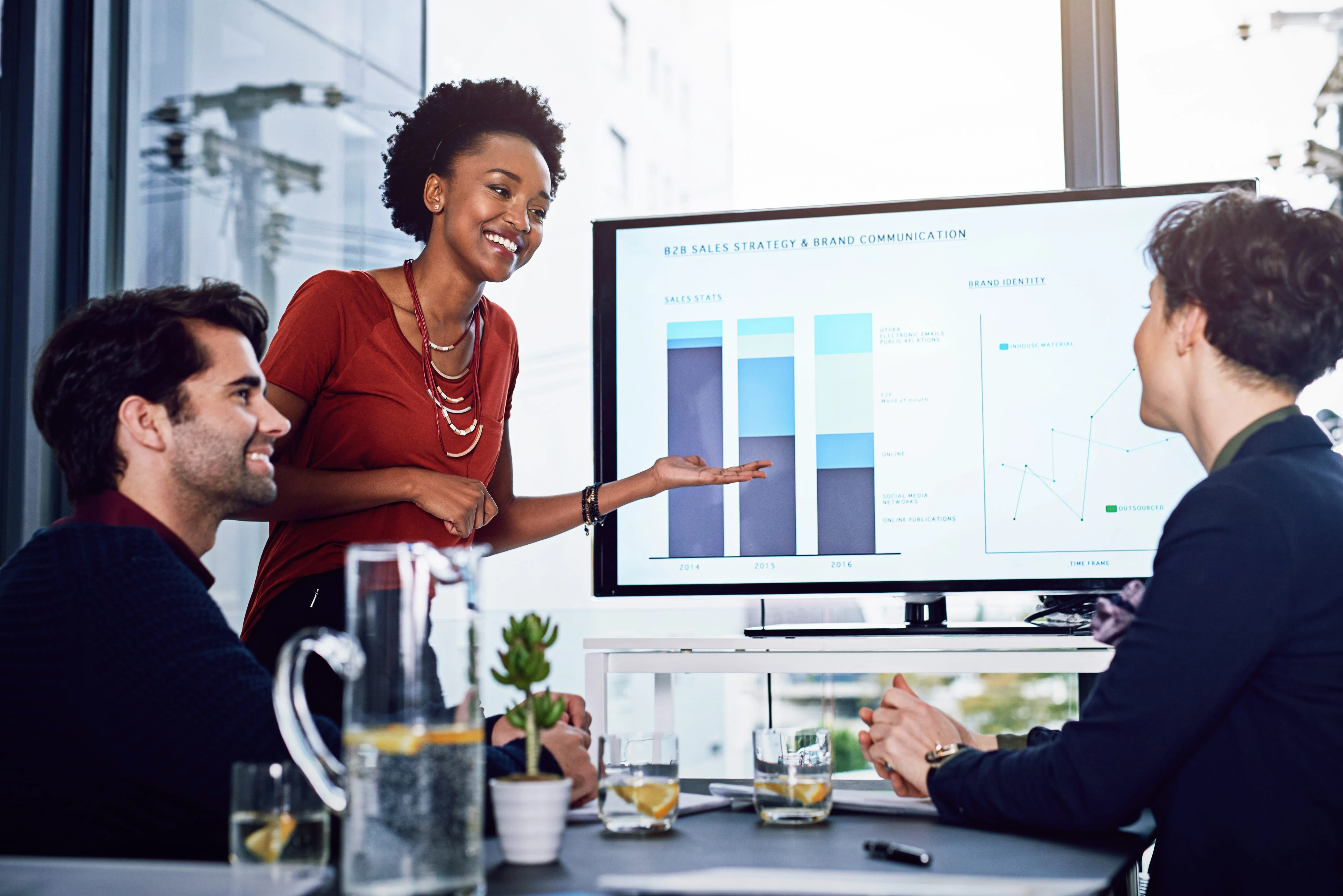 Three professionals in a meeting room with a woman presenting sales and brand communication charts on a large screen.
