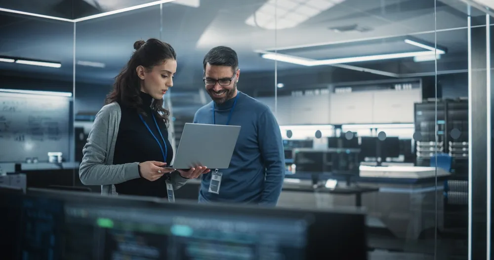 Two professionals in an office looking at a laptop together, surrounded by computer equipment.