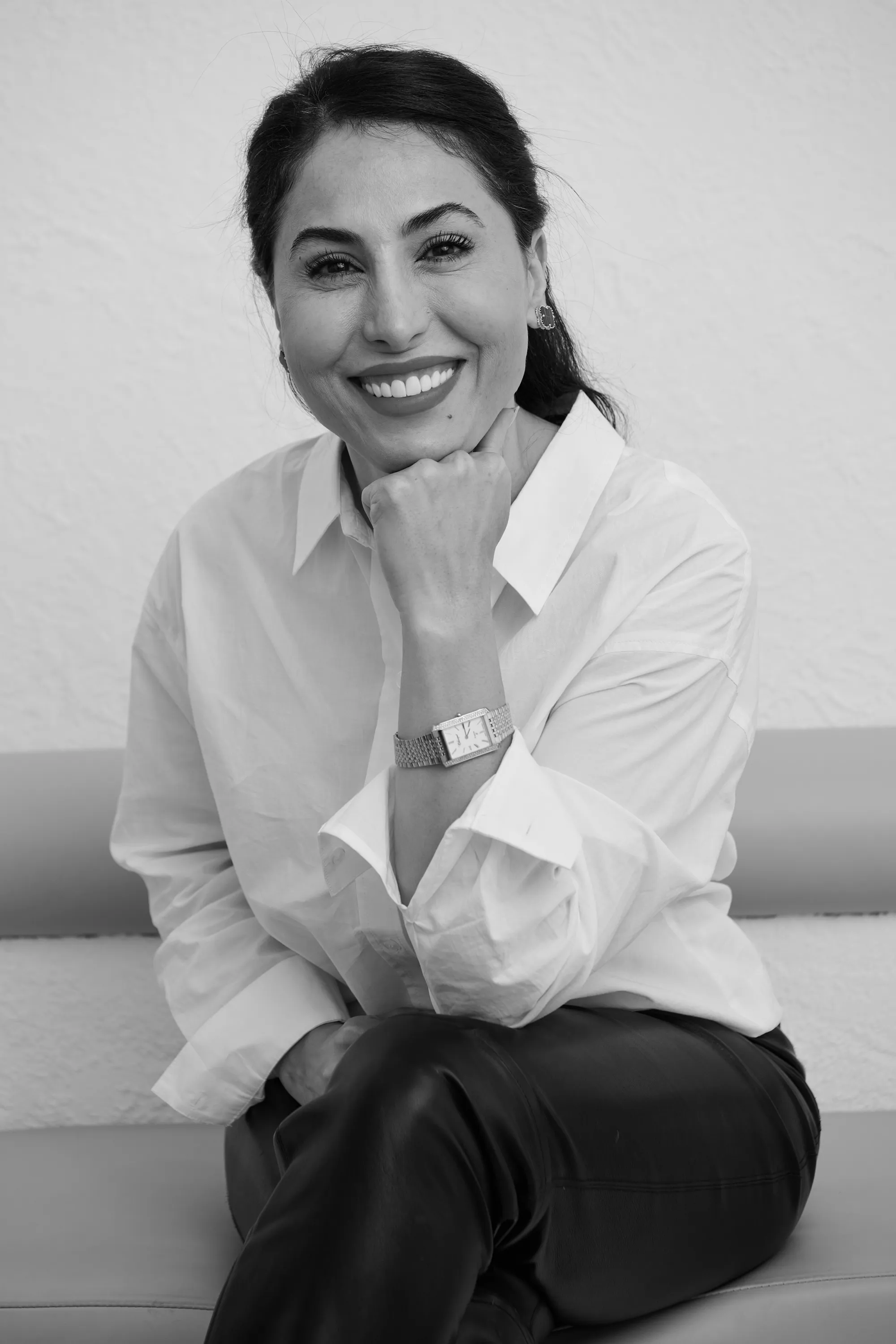Smiling woman with dark hair wearing a white blouse and leather pants, sitting with her chin resting on her fist.