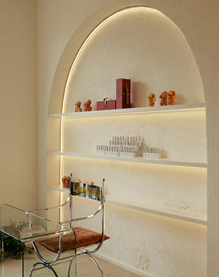 Minimalist corner with three white illuminated shelves in an arched wall niche, displaying small sculptural candles, boxed products, and cosmetic bottles, with a modern chrome and glass chair with a brown cushion in front.