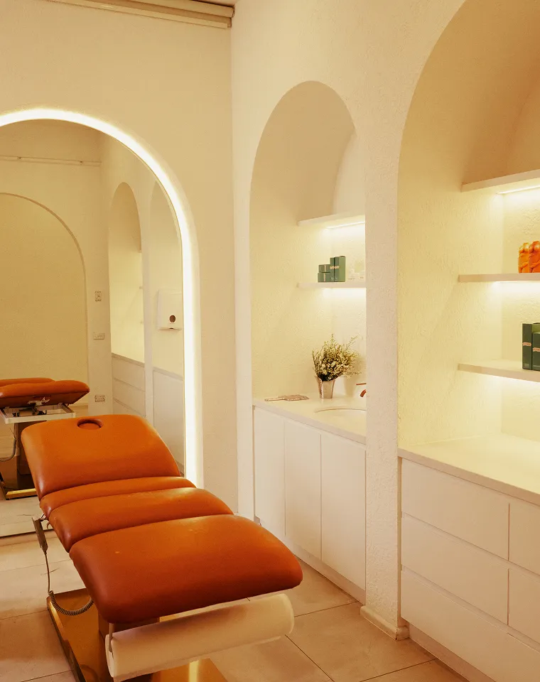 Minimalist spa or treatment room with a brown cushioned reclining chair, arched wall niches holding shelves with products, and soft warm lighting.