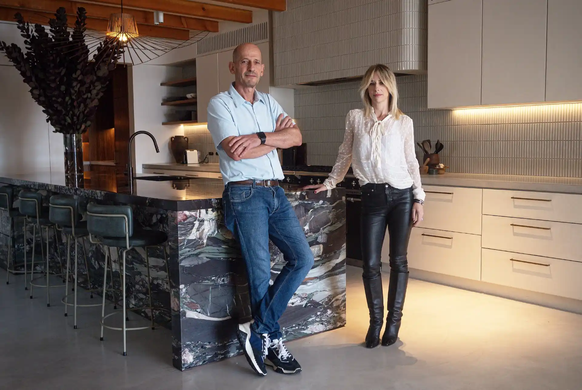 Man and woman standing in a modern kitchen with a marble island and bar stools, under warm lighting.