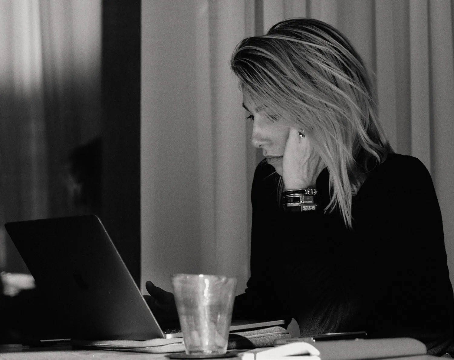 Black and white photo of a woman with shoulder-length hair focused on a laptop, resting her head on one hand.
