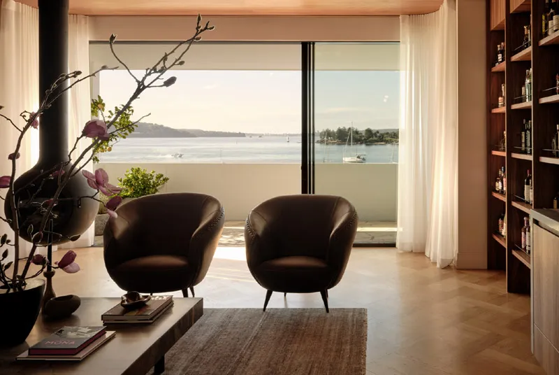 Cozy living room with two dark armchairs facing large glass doors showing a waterfront view with sailboats.