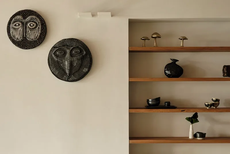 Minimalist wooden shelves mounted on a beige wall holding ceramic mushrooms, bowls, vases, and decorative items, alongside two circular tribal-style wall masks.