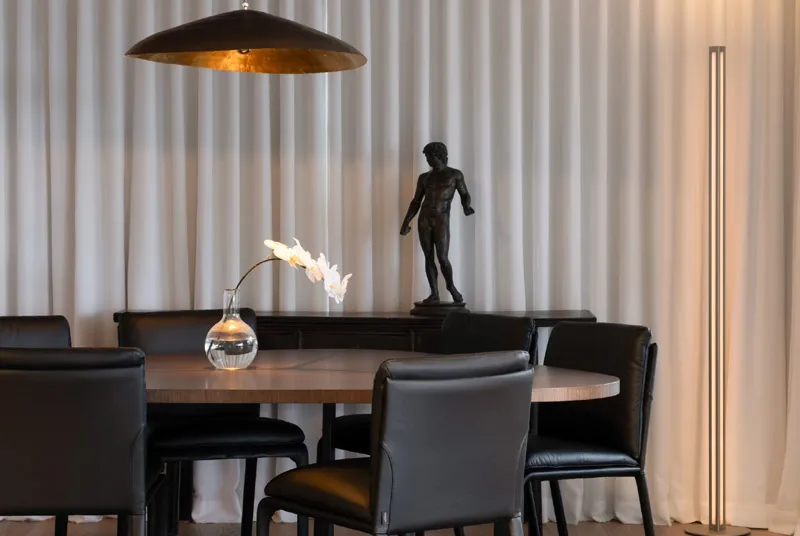 Modern dining room with black leather chairs around a wooden table, a glass vase with white flowers, a dark statue on a sideboard, and warm pendant and floor lighting.