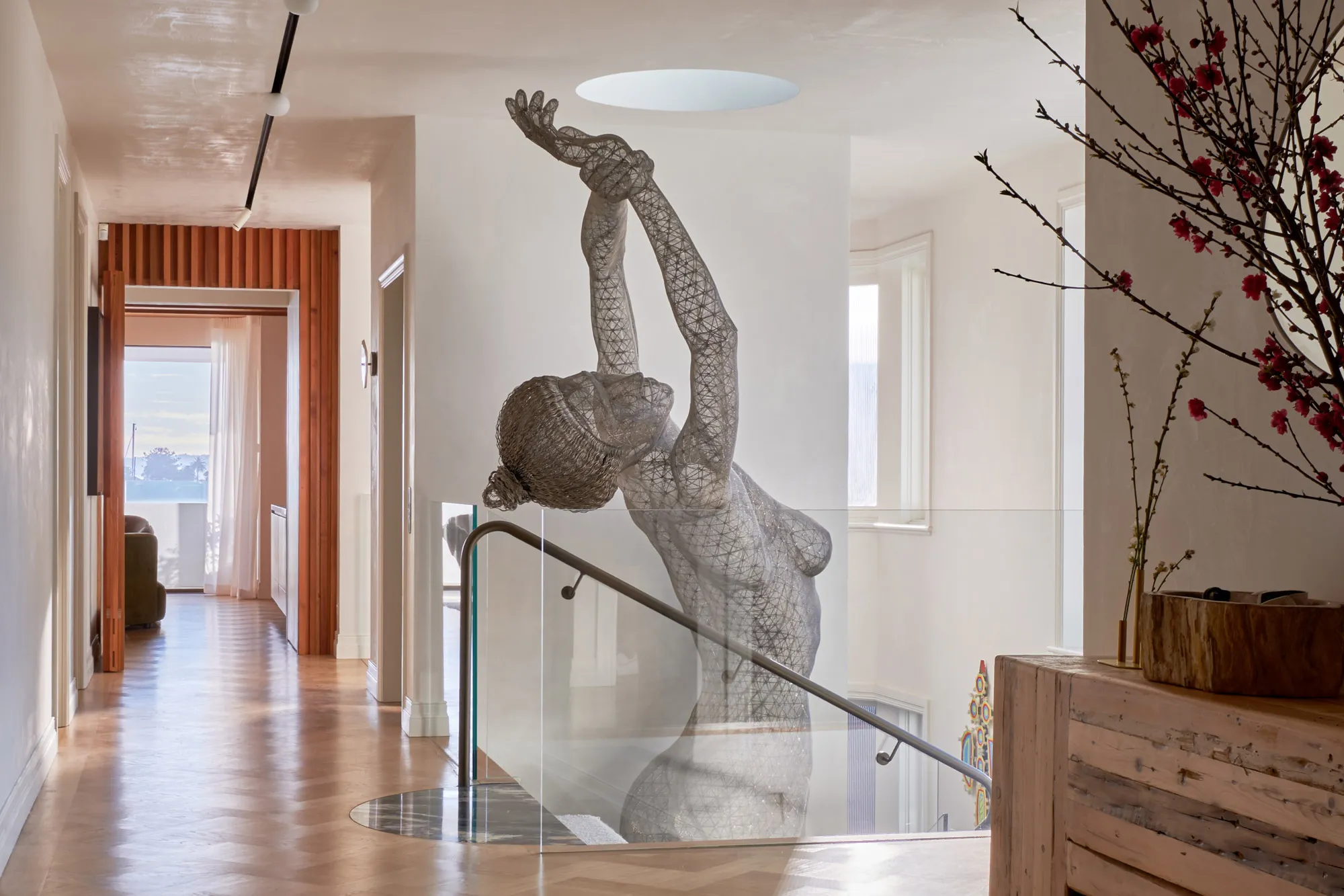 Wireframe sculpture of a woman with arms raised in a bright modern interior next to a staircase.