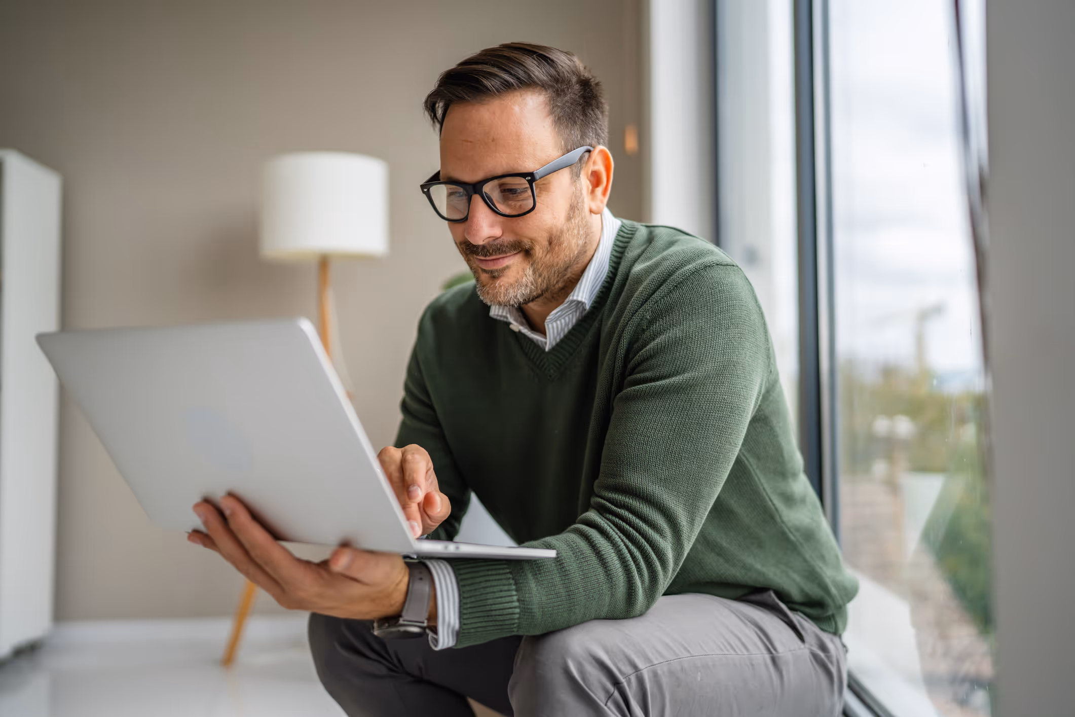 Man wearing glasses and green sweater sitting by a window using a laptop.