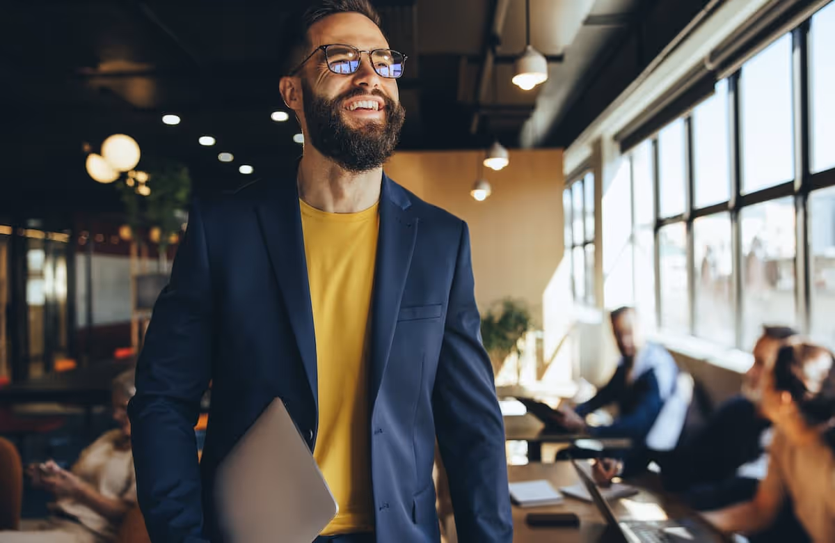 Smiling bearded man wearing glasses and a navy blazer holding a laptop in a modern office with people working in the background.