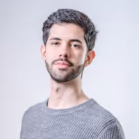 Young man with short curly dark hair and a trimmed beard wearing a gray sweater against a light gray background.