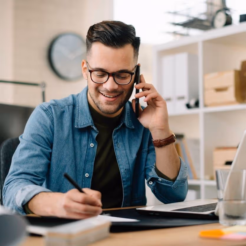 Smiling man wearing glasses and denim shirt talking on phone while writing at desk with laptop and papers.