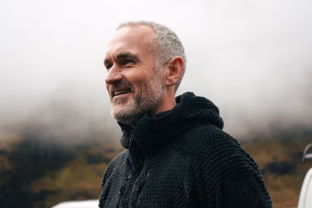 Smiling middle-aged man with gray hair and beard wearing a black textured jacket outdoors with a foggy background.