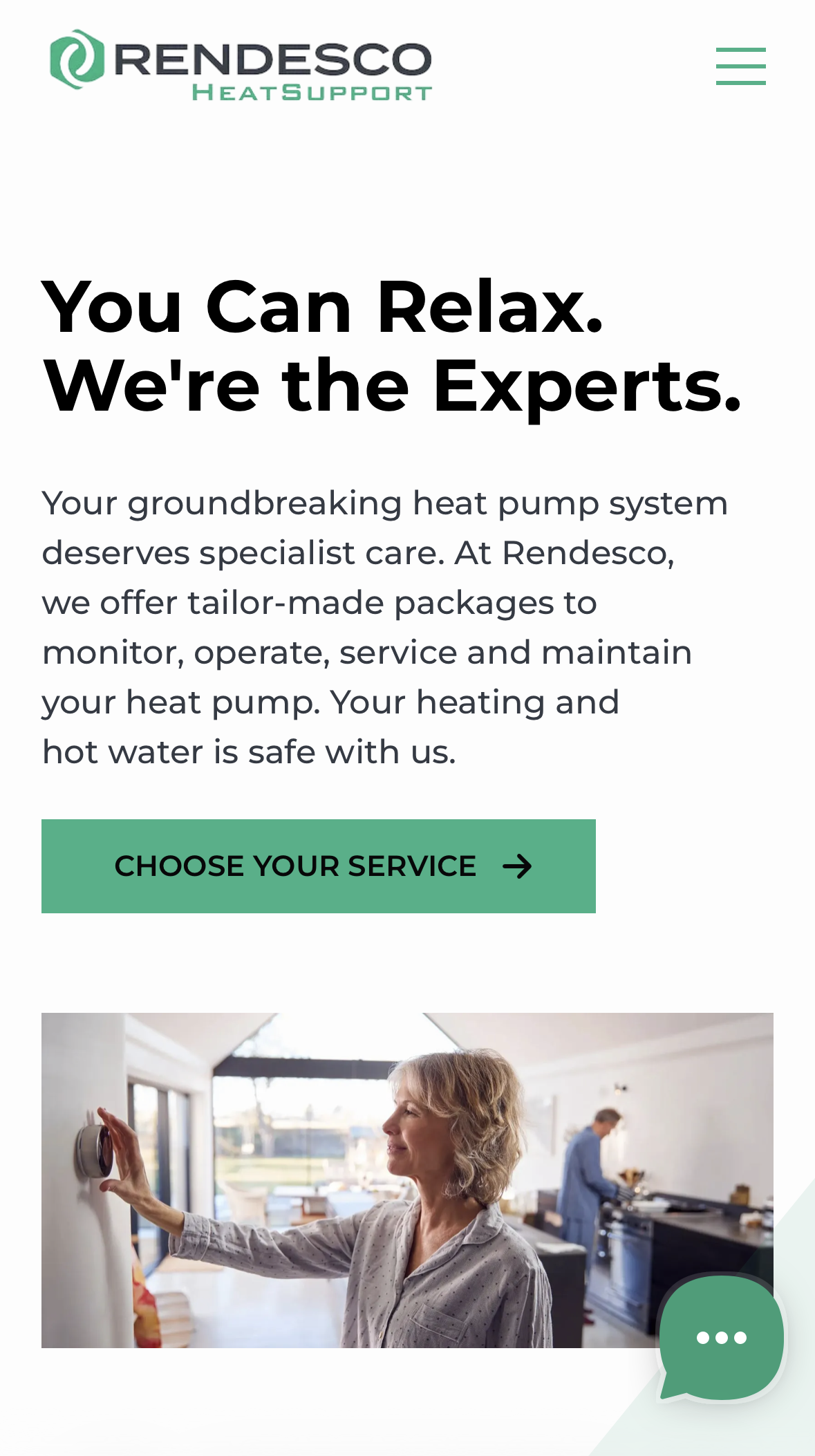 Middle-aged woman adjusting a wall thermostat in a modern kitchen while a man cooks in the background.