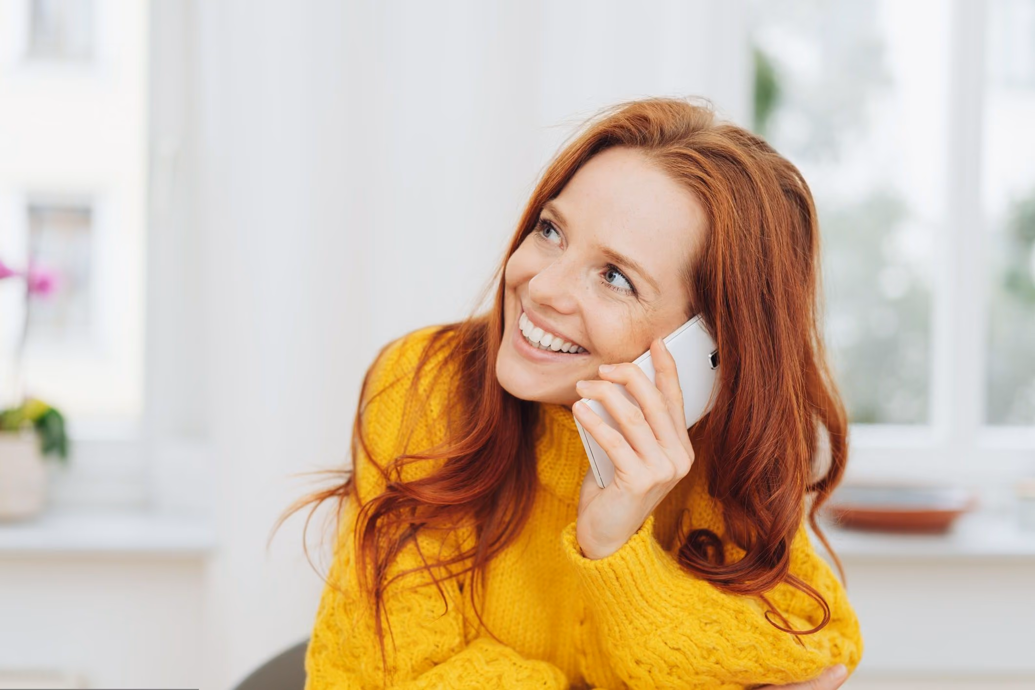 Smiling red-haired woman in a yellow sweater holding a white smartphone to her ear indoors.