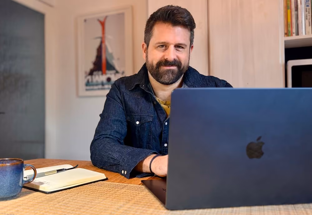 Man with beard and dark shirt sitting at table using laptop with mug and notebook beside him.