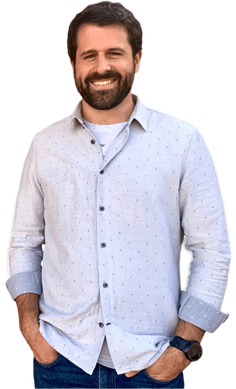 Smiling man with dark hair and beard wearing a light blue patterned button-up shirt and jeans, hands in pockets.