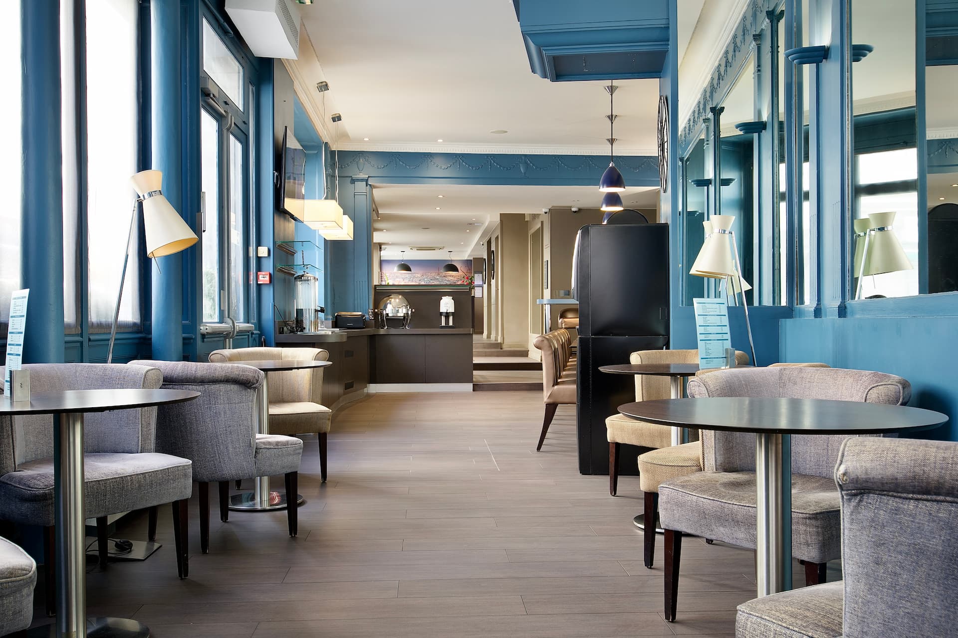Salon d'hôtel élégant avec murs bleus, fauteuils gris et beiges autour de tables rondes, lampes sur pieds, et buffet au fond.