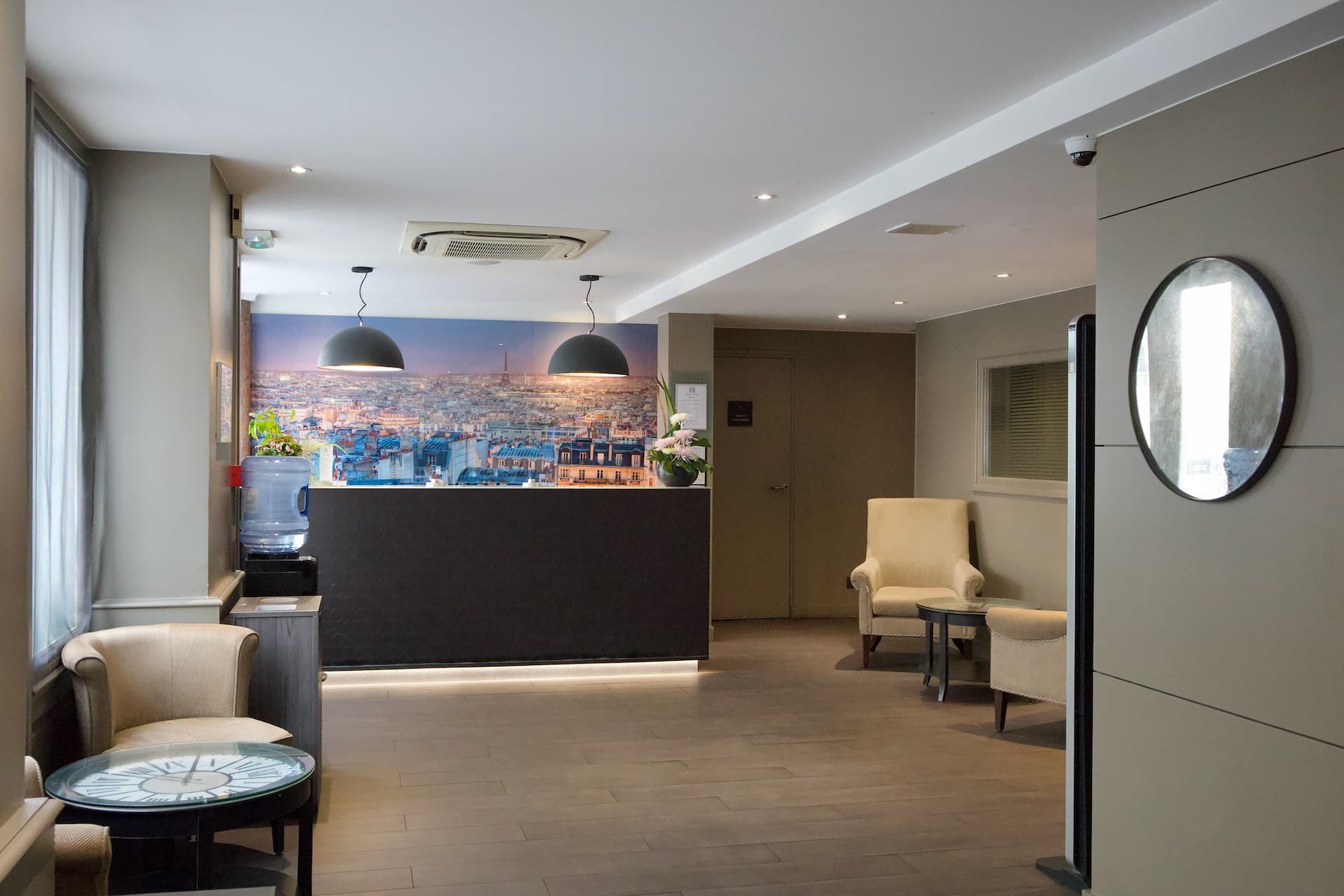 Lobby d'hôtel moderne avec comptoir noir, deux fauteuils beige, lampe suspendue et une grande photo de Paris avec la Tour Eiffel en arrière-plan.