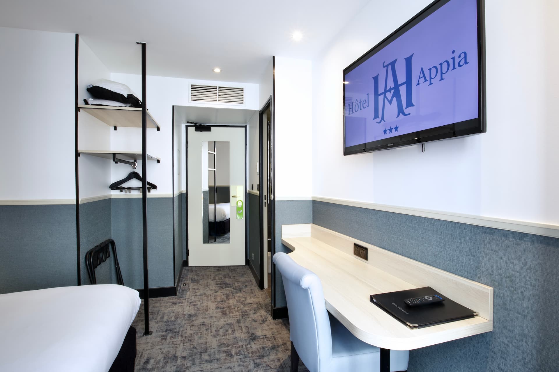 Chambre confort avec lit blanc, bureau en bois clair, chaise bleue, télévision murale affichant Hôtel Appia, et porte avec miroir.