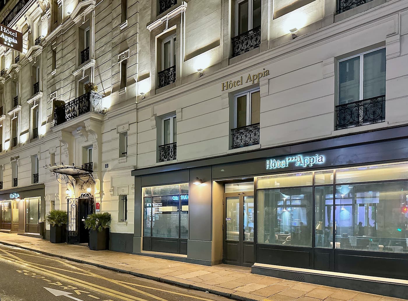 Façade éclairée de l'Hôtel Appia avec fenêtres à balcons en fer forgé et entrée avec auvents en fer noir.