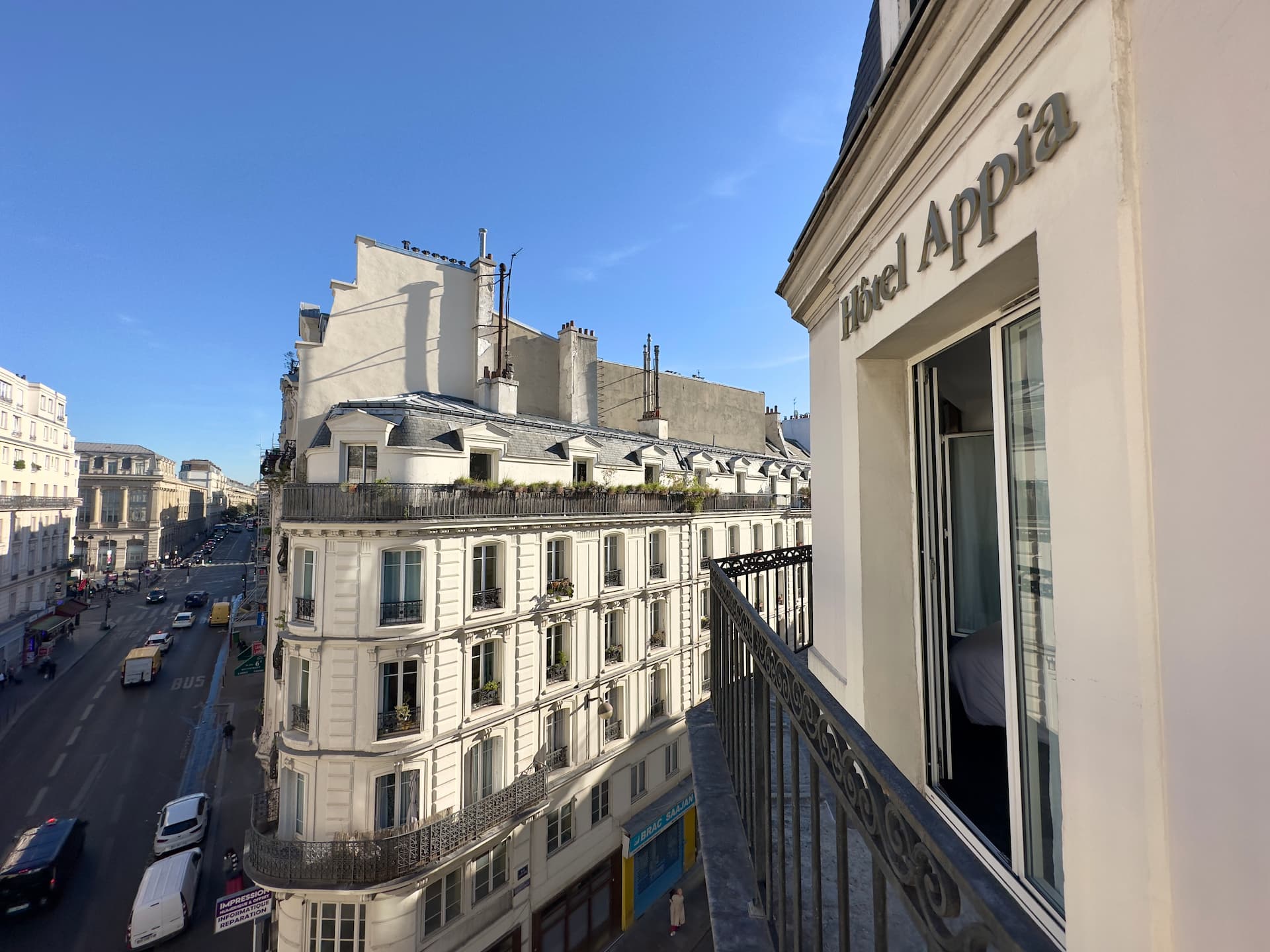 Vue depuis un balcon de l'Hôtel Appia montrant un immeuble haussmannien et une rue animée sous un ciel bleu clair.