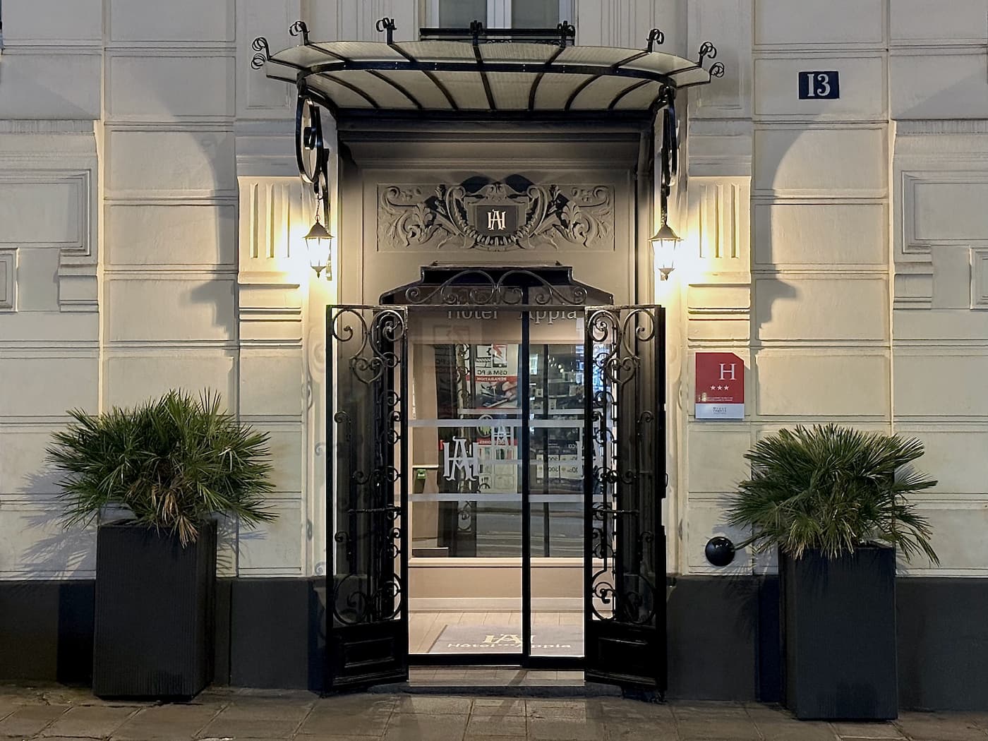 Entrée d'hôtel élégante avec une porte en verre encadrée de métal noir orné, deux grandes plantes en pot et deux lanternes allumées de chaque côté.