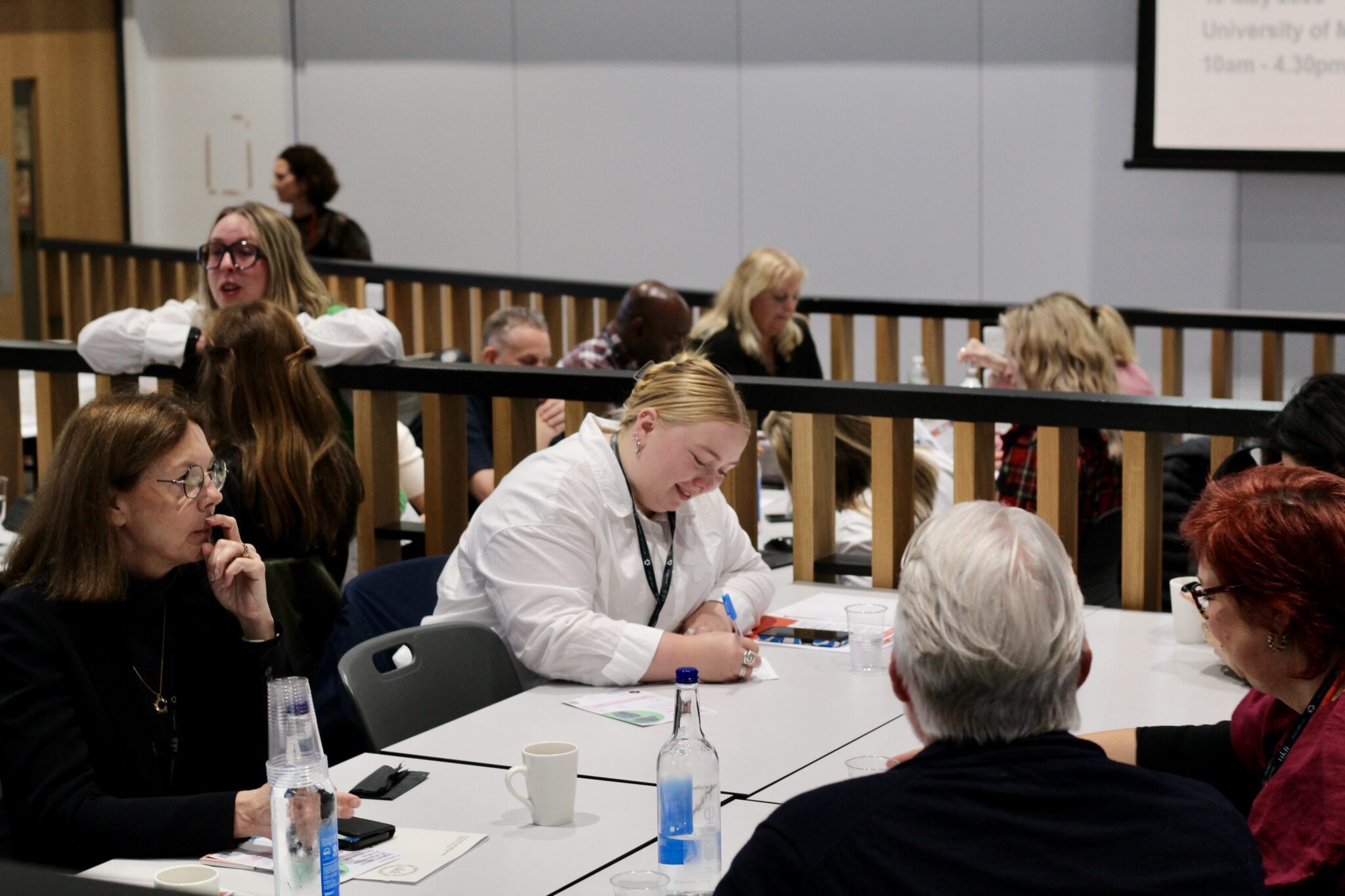 People sitting at tables in a conference or meeting room, engaging in discussion and note-taking.