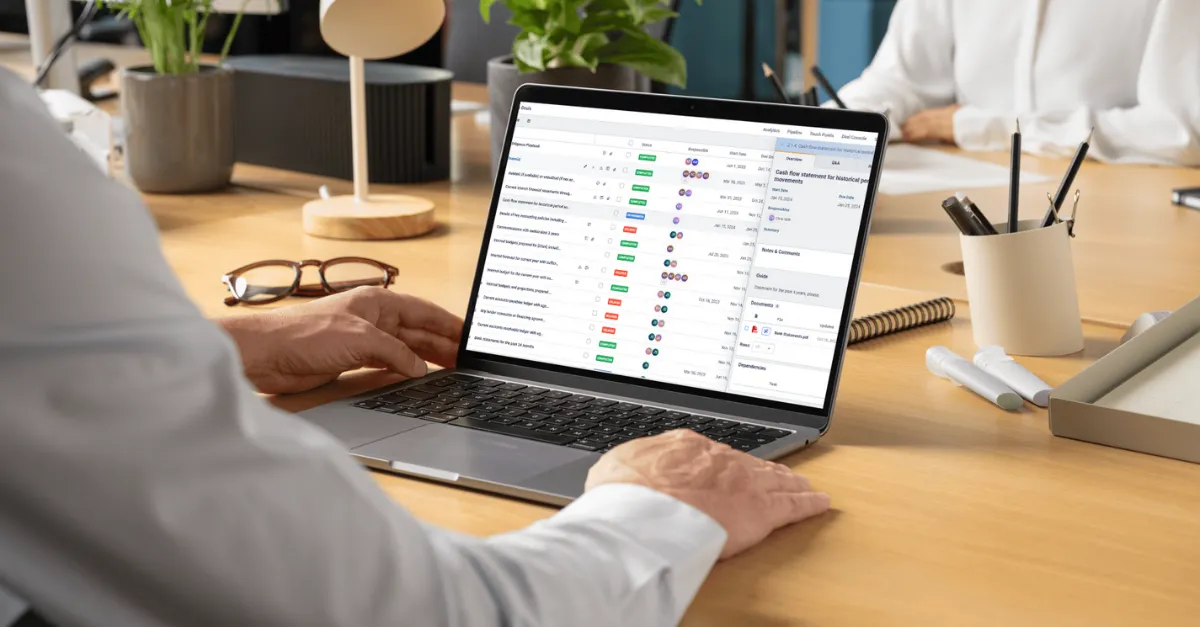 Person working on a laptop at a wooden desk showing a deals management dashboard with task statuses and responsible team members.