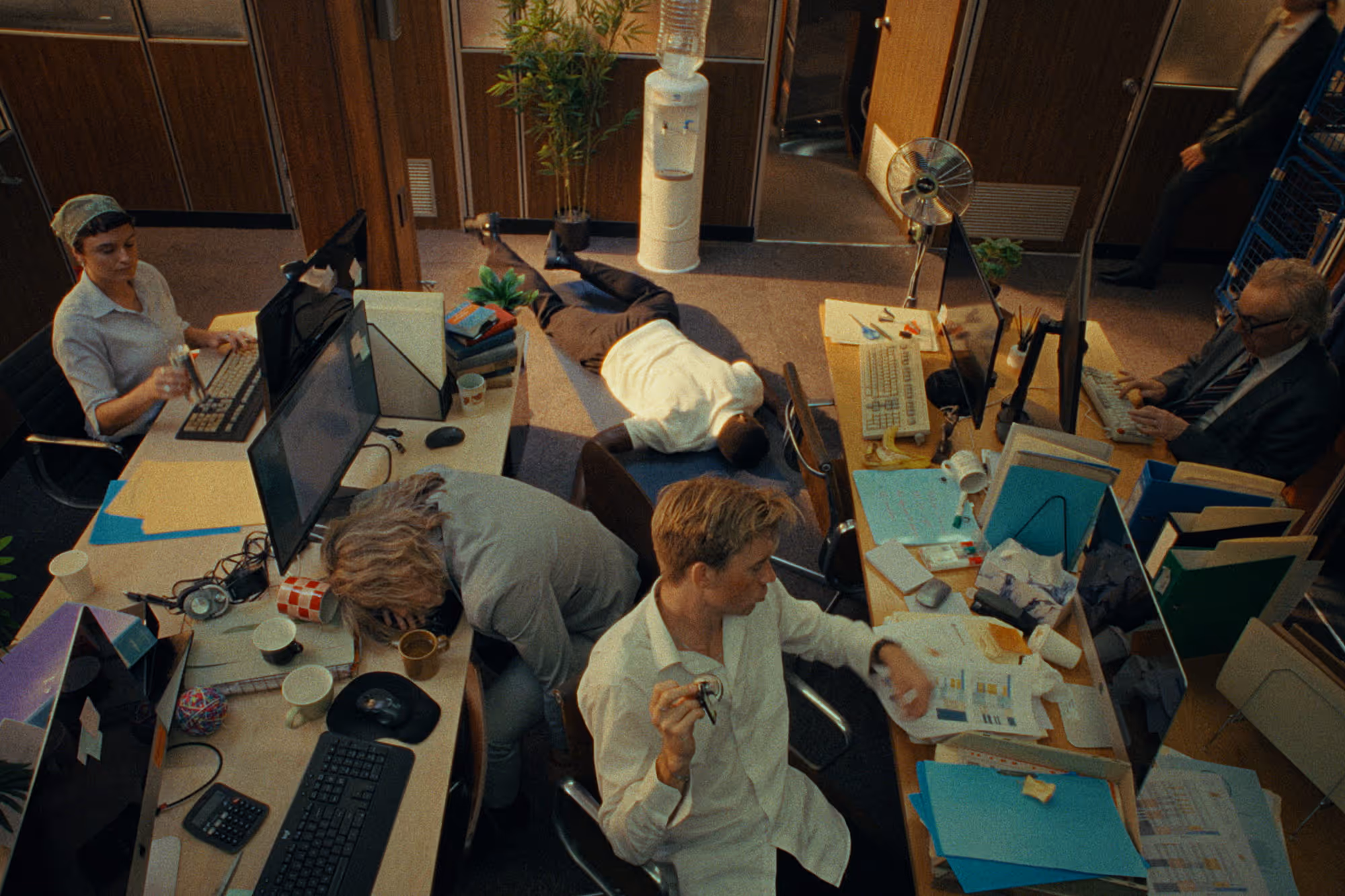 Office scene with several people appearing exhausted or passed out around cluttered desks with computers and paperwork.