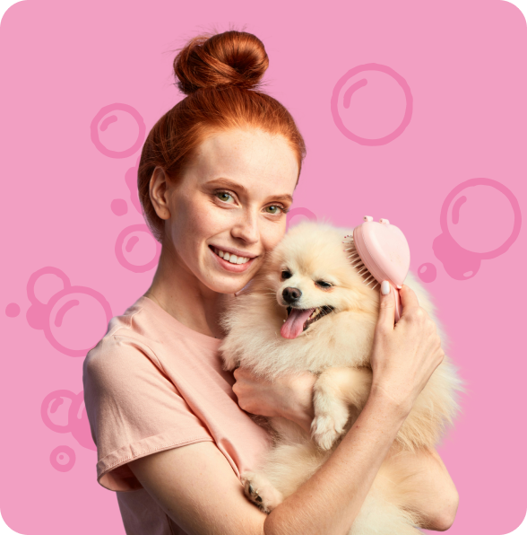 Smiling woman with red hair in a bun holding and brushing a happy fluffy white dog against a pink background with bubble illustrations.