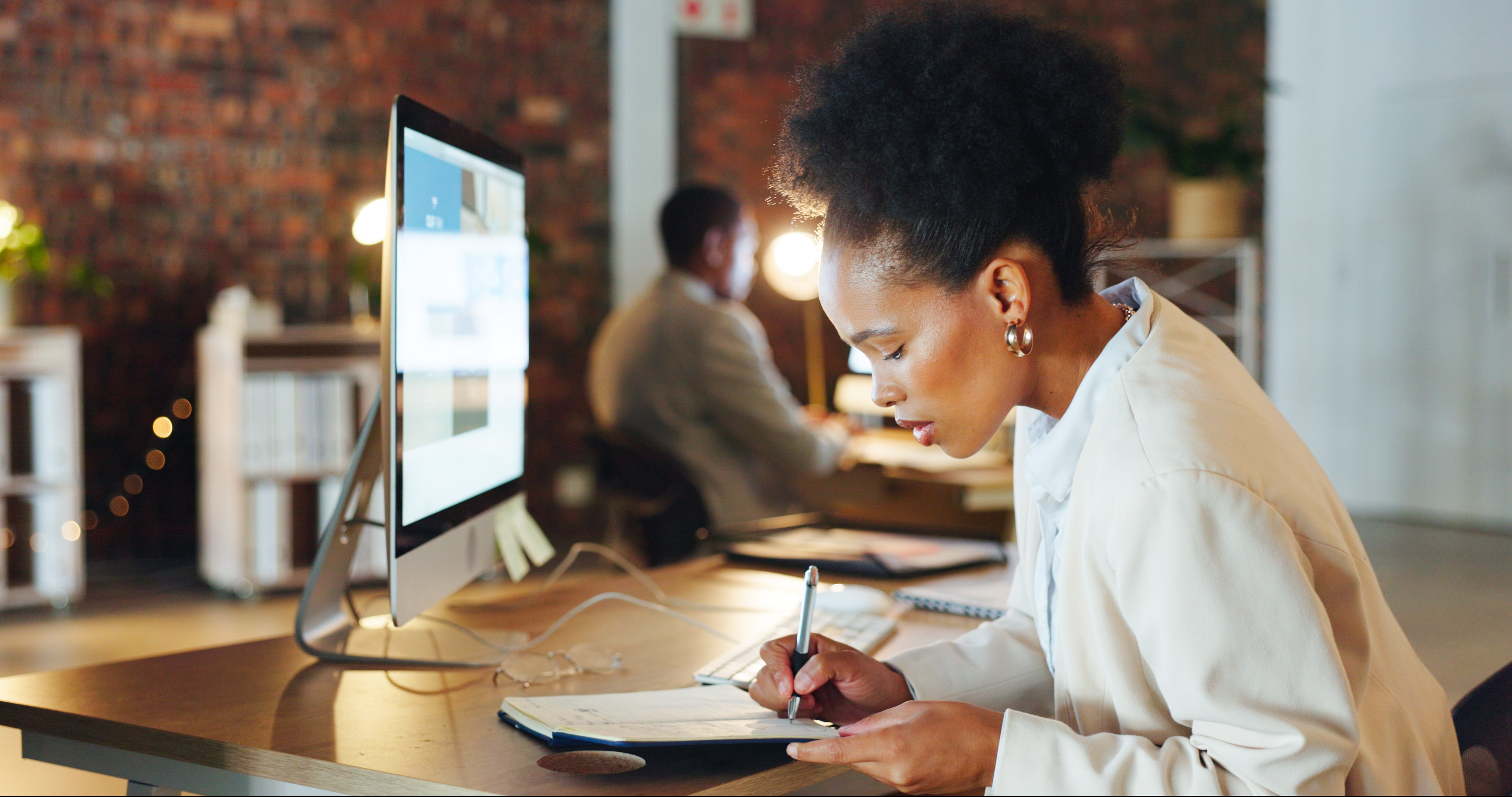 Professional woman working at a desk in a modern office, writing notes on a clipboard beside a desktop computer, with a colleague working in the background.