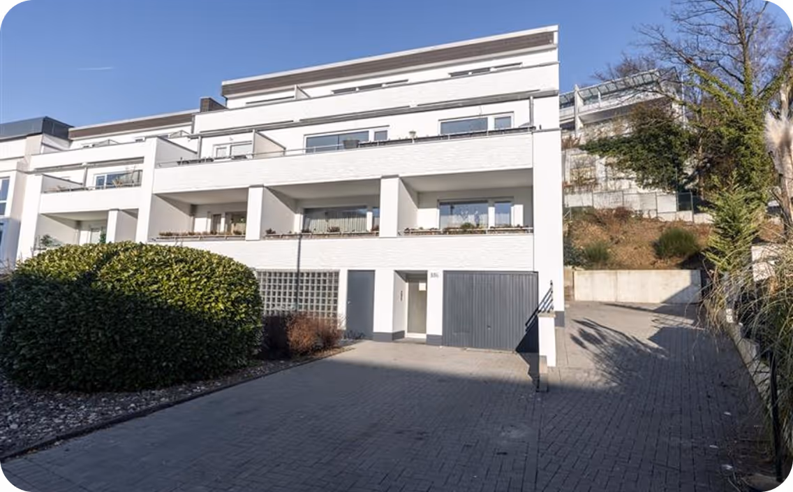 Modern white multi-family house with balconies, garage, and paved driveway under a clear blue sky.