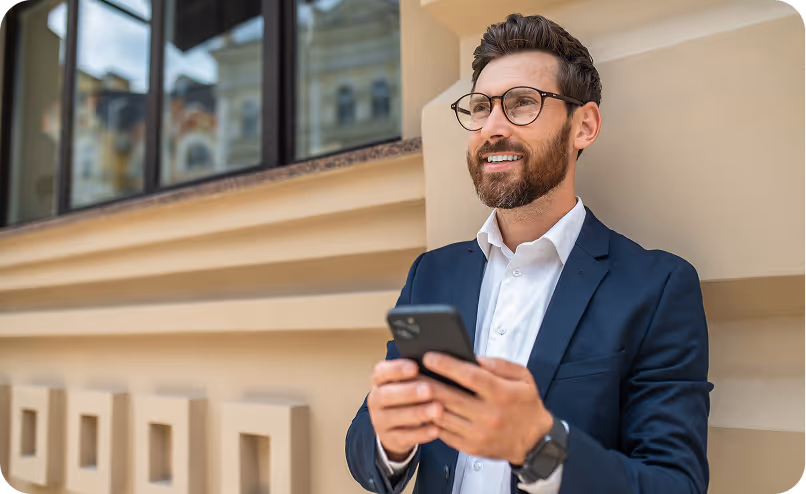 Smiling man in a navy suit and glasses holding a smartphone outdoors near a beige building.